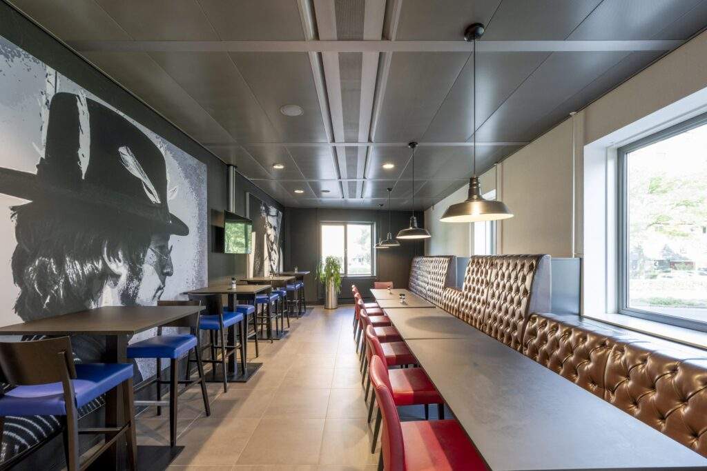 Modern ingerichte eet- of werkruimte aan het Koningin Wilhelminaplein met lange tafels, gekleurde stoelen, kunst aan de muur en grote ramen.